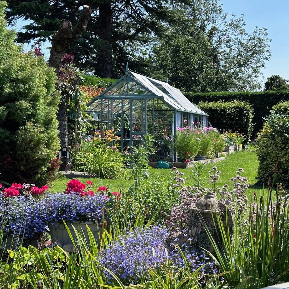 A glass Rhino Greenhouse stands amidst vibrant flower beds with colorful blooms, surrounded by lush greenery and tall trees, reflecting a sunny garden scene.
