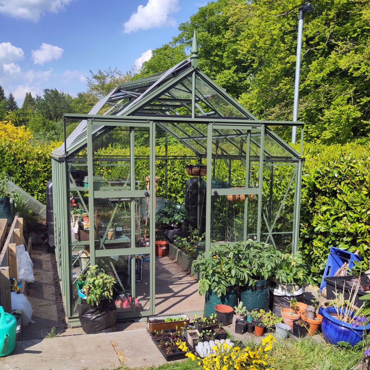 A green Rhino Greenhouse stands amidst vibrant plants, with gardening tools and pots scattered around in a lush garden setting under a partly cloudy sky.