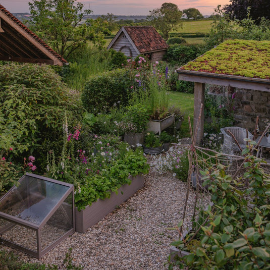 A small Rhino Greenhouse sits in a lush garden with blooming flowers and greenery, surrounded by gravel pathways. Nearby, wooden sheds with red-tiled roofs blend into the peaceful rural landscape.