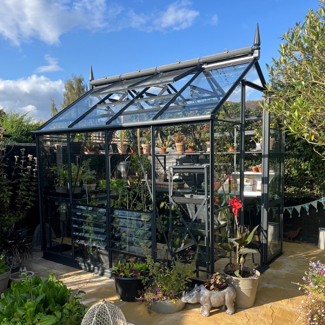A glass Rhino Greenhouse containing various potted plants stands on a sunlit patio, surrounded by greenery and decorative garden items, under a blue sky with scattered clouds.