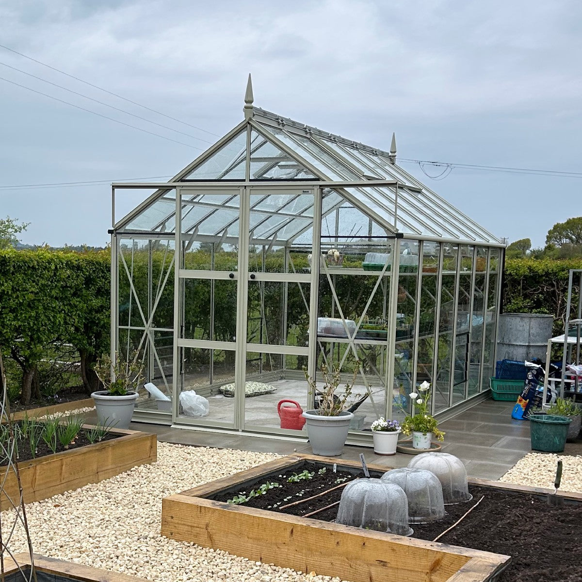 A glass Rhino Greenhouse stands in a garden, surrounded by raised wooden beds and gravel paths. Potted plants and gardening tools are visible nearby, creating a well-maintained planting area.