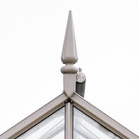 A pointed metal finial tops the intersecting edges of a glass Rhino Greenhouse roof. The finial is prominently displayed against a plain white background, emphasizing architectural detail.