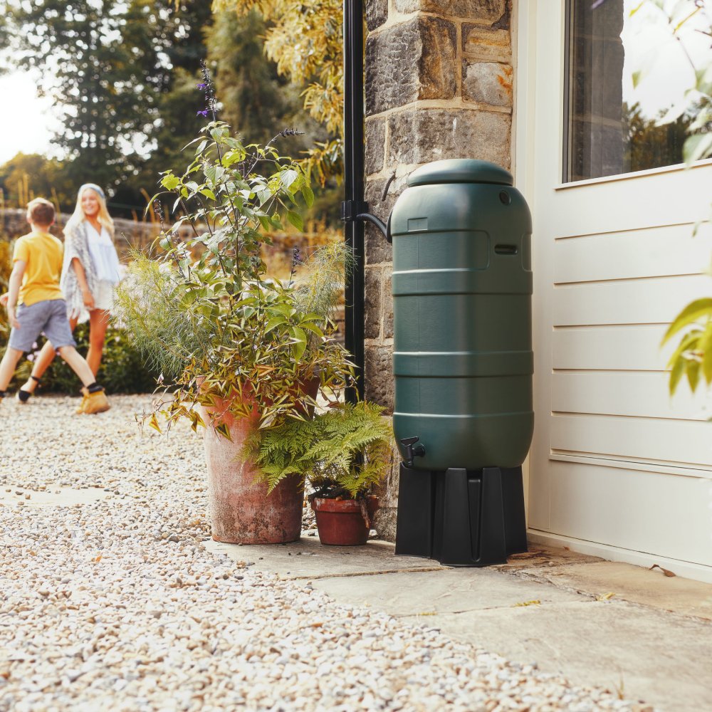 A green rain barrel stands next to a stone building, surrounded by potted plants on a gravel path. In the background, two people walk along the pathway.