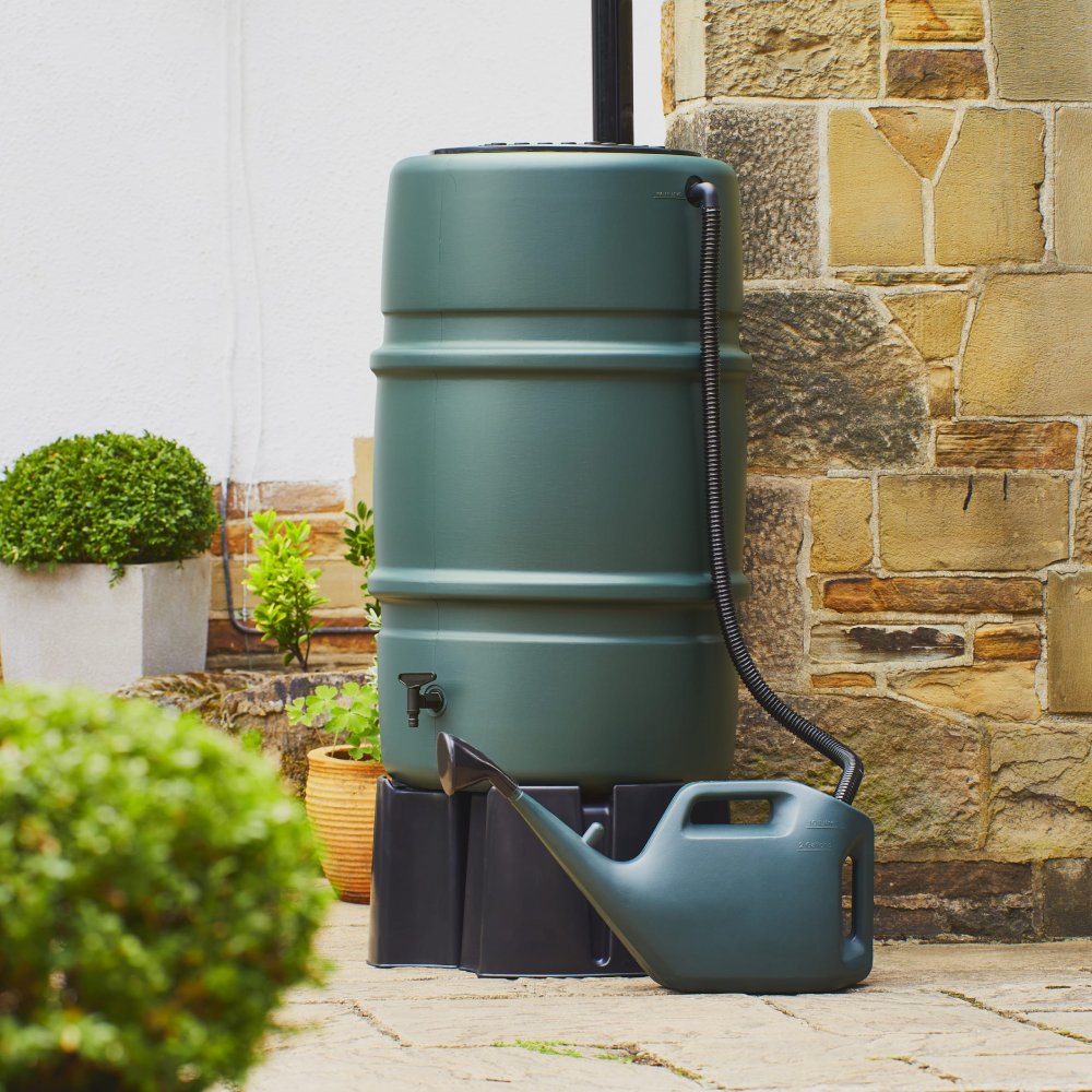 A large green water butt stands next to a stone wall, connected to a downspout. In front, a matching green watering can rests on the paved ground, surrounded by potted plants.