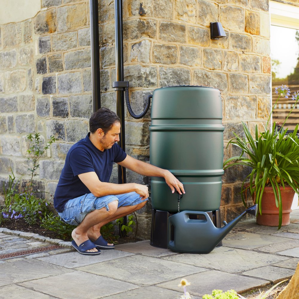 A large green rainwater barrel, fitted with a spigot and hose, stands beside a brick wall. A green watering can rests beneath, surrounded by potted plants in a garden setting.