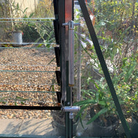 Metallic spring mechanism adjusting glass louver windows in a Rhino Greenhouse, surrounded by lush plants and gravel-filled ground, with sunlight filtering through the structure.