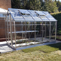 A glass Rhino Greenhouse with open windows sits on a paved area, surrounded by grass and trees, with some plants growing inside.