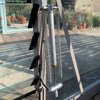 A metal Rhino Greenhouse window opener with springs is partially open, revealing potted plants and paving stones outside. Sunlight creates shadows on the transparent panels.