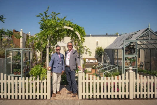 Two people stand in front of a white picket fence and lush greenery with a Rhino Greenhouse visible behind. A sign reads Rhino in the background, under a clear blue sky.