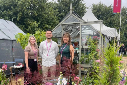 Three people stand smiling in front of a display of Rhino Greenhouses surrounded by vibrant plants and flowers. They're wearing name tags, set against a backdrop of green foliage and promotional banners.