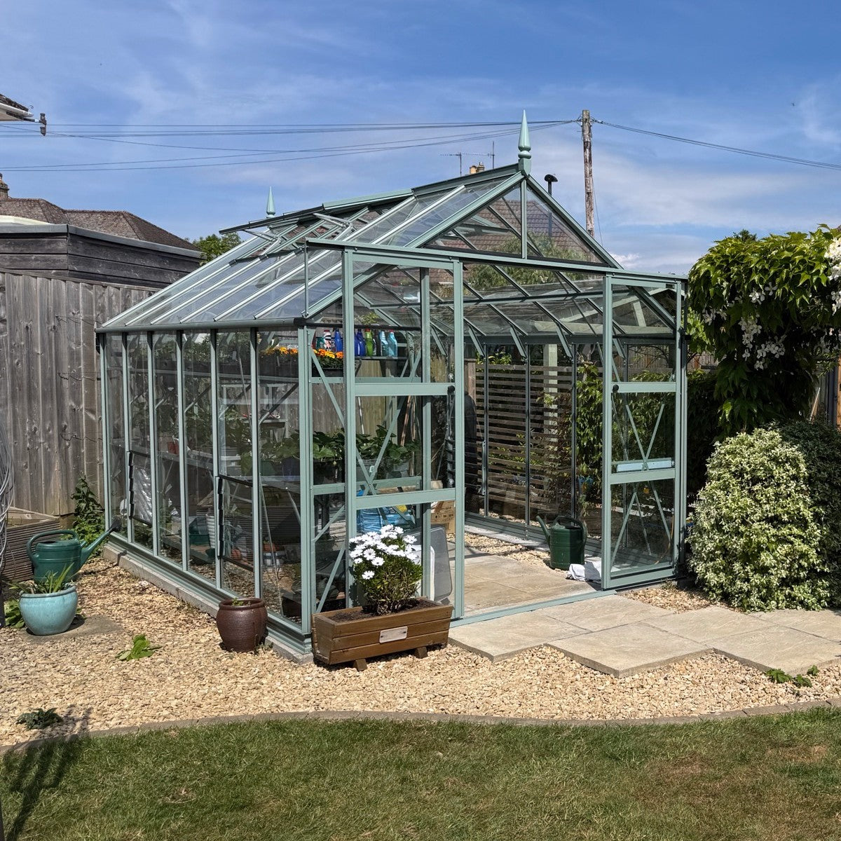 A glass Rhino Greenhouse stands in a garden, surrounded by gravel and potted plants. Inside, there are visible plants and gardening tools. It's a bright day with clear blue skies.
