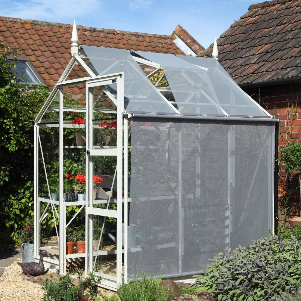 A white-framed Rhino Greenhouse with an open door displays potted plants inside. It is situated in a garden area, surrounded by bushes and adjoining a brick building with tiled roofs.