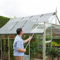 Man adjusting retractable shade on a glass Rhino Greenhouse using a pole; interior filled with thriving plants and gardening supplies, set in a lush garden environment.