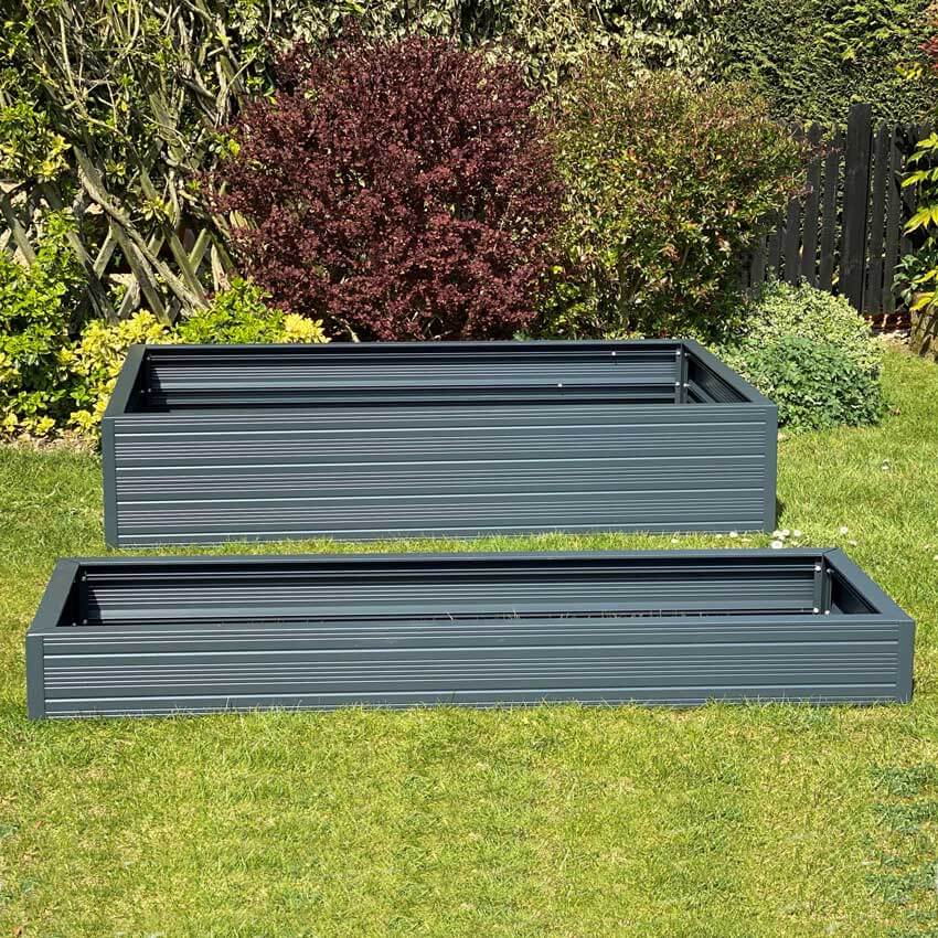 Two rectangular, metal garden planters rest on a grassy lawn. The background features lush greenery, including various shrubs and trees, creating a garden setting.